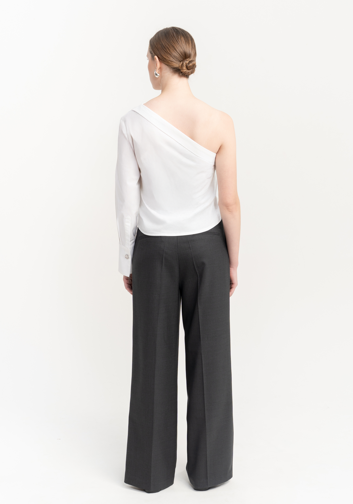 Woman wearing a white one-shoulder top and gray wide-leg trousers on a white background - back view