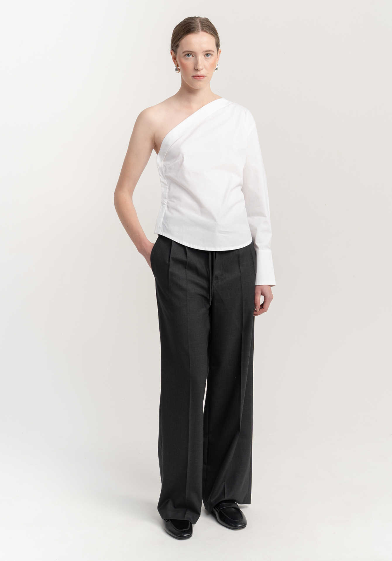 Woman wearing a white one-shoulder top and black trousers on a light gray background - front view